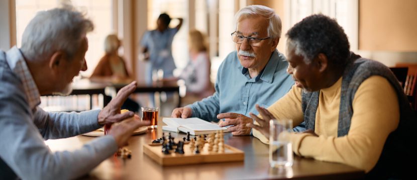 Mature man reading book and communicating with male friends while they are playing chess at retirement community.