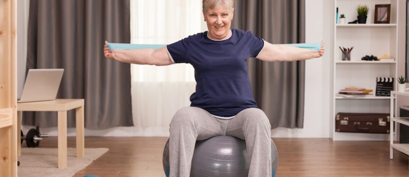 Senior woman working out at home with fitness bands to solve her health problems. Old person pensioner online internet exercise training at home sport activity with dumbbell, resistance band, swiss ball at elderly retirement age