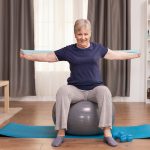 Senior woman working out at home with fitness bands to solve her health problems. Old person pensioner online internet exercise training at home sport activity with dumbbell, resistance band, swiss ball at elderly retirement age